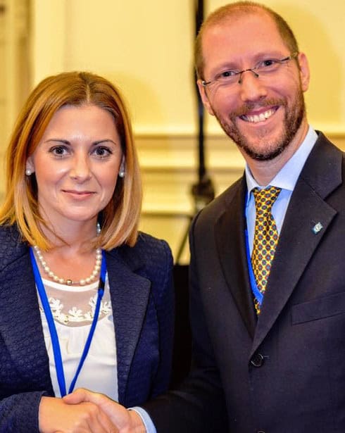 Humberto, indossando un abito blu, una camicia celeste e una cravatta gialla con vari fiori rossi e blu, stringe la mano con il Segretario Parlamentare maltese, una donna dalla pelle chiara e capelli biondi, indossante una giacca blu, maglietta bianca elegante in pizzo, e una collana e orecchini di perle. Entrambi stanno sorridendo