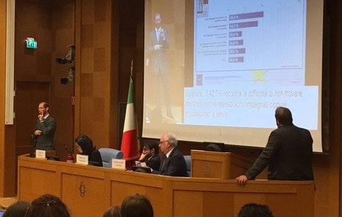 Immagine in un'aula del parlamento italiano. Humberto è alla tua sinistra, in piedi, mentre fa una relazione in lingua dei segni italiana. Alla sua destra c'è una cattedra con alcuni esperti seduti che lo ascoltano attentamente. Uno di loro guarda direttamente il grande schermo alle spalle di Humberto, dove è proiettata una slide in collegamento alla sua relazione.