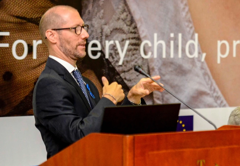 Humberto è in piedi, indossa un abito blu, una camicia bianca e una cravatta blu, mentre presenta una relazione sulla UNICEF. Alle sue spalle, è visibile un grande cartellone con la scritta 'For every child, protection and...