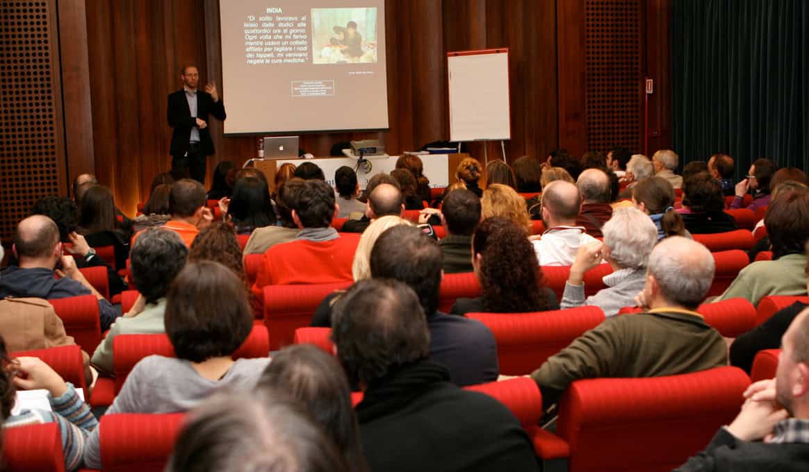 Humberto è in piedi all'estremità della stanza, di fronte al pubblico, mentre tiene una lezione. Accanto a lui, si trovano un videoproiettore e una lavagna con fogli bianchi. Di fronte a Humberto, sulle poltrone rosse, sono visibili le teste di centinaia di persone sedute