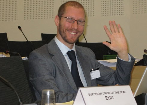 Humberto è seduto, saluta con la mano, e sul tavolo c'è un cartellone con la scritta 'European Union of the Deaf - EUD’