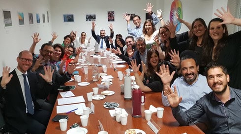 Foto di gruppo con circa quaranta responsabili dei vari ministeri del governo argentino e rappresentanti di associazioni delle persone con disabilità, seduti attorno a un lungo tavolo. Sul tavolo sono presenti vari fogli e penne, bicchieri di carta, un termos per l'acqua calda, bustine di tè, una caraffa d'acqua e biscottini, facturas (varietà di croissants tipici argentini). Sui muri sono affissi vari fogli che illustrano le battaglie condotte dall'INADI, e una grande insegna 'INADI' con il logo a forma di sole è visibile. Tutti sorridono e applaudono con le mani in aria, seguendo il gesto simbolico dell'applauso nella cultura sorda