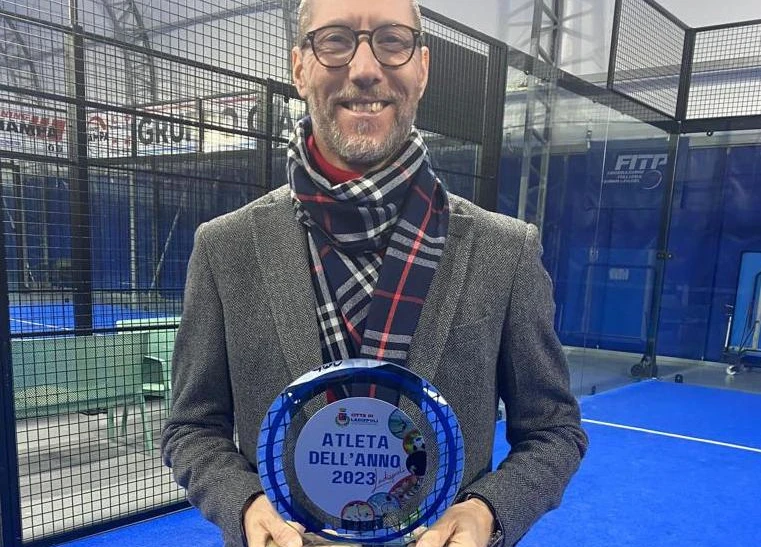 Humberto è ritratto in piedi, sorridente, indossa una giacca grigia e una sciarpa a quadretti blu, rosso e bianco. Si trova all'interno di un campo da padel e tiene in mano una targa con un cerchio in metallo e una cornice esterna di colore blu elettrico. Sulla targa è scritto 'Città di Ladispoli, atleta dell'anno 2023'.