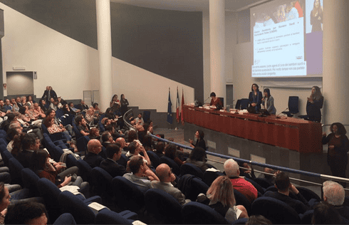 Immagine dei partecipanti seduti, concentrati nell'ascoltare una relazione in una grande aula universitaria. La scena comprende diversi interpreti: lingua dei segni italiana, lingua dei segni tattile italiana, segni internazionali e sottotitoli