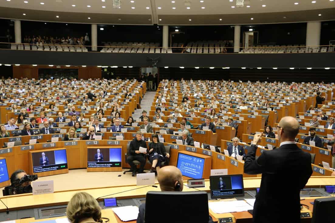 Humberto Insolera si trova sul palco dell'Emicidio al Parlamento europeo, moderando un discorso con la lingua dei segni. Di fronte a lui, il Parlamento europeo è visibile, con oltre 700 persone che lo osservano attentamente. Un giornalista è presente, focalizzato su Humberto, mentre lo filma con la sua telecamera