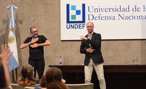 Humberto, indossando una giacca blu, una camicia a righe bianco e celeste e pantaloni color crema, è in piedi mentre spiega il suo lavoro su EDF utilizzando la lingua dei segni argentina (LSA). Alla sua destra si trova una bandiera dell'Argentina e un'interprete che traduce dalla LSA a segni internazionali. Alle sue spalle è visibile un'insegna con la scritta 'UNDEF - Universidad de la Defensa Nacional'. Davanti a lui, alcune teste delle persone sono visibili mentre lo stanno guardando