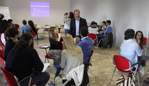 Immagine rappresentata in una stanza chiusa, dove ci sono cinque gruppi di giovani divisi in cinque, impegnati in discussioni durante il workshop. Un gruppo sta chiedendo informazioni a Humberto. In fondo, è visibile una diapositiva con la scritta 'Gruppi di lavoro / Simulazioni', mentre una persona sta registrando la scena con una telecamera