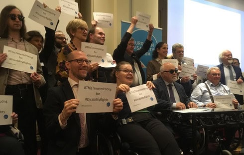 Immagine di un gruppo misto di membri dell'EDF, alcuni seduti e altri in piedi, mentre mostrano un foglio con la scritta 'hashtag ThisTimeImVoting for hashtag DisabilityRights'. Humberto è presente in basso, sulla tua sinistra, sorride mentre tiene il foglio tra le mani.