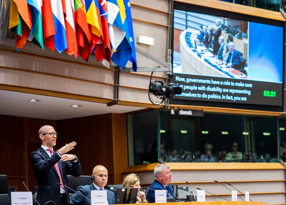 Humberto Insolera si trova in piedi sul palco del Parlamento europeo, impegnato in un discorso in lingua dei segni. Alle sue spalle, a sinistra, sono esposte le bandiere di vari paesi europei, mentre a destra c'è un monitor che proietta un video con sottotitoli in lingua inglese