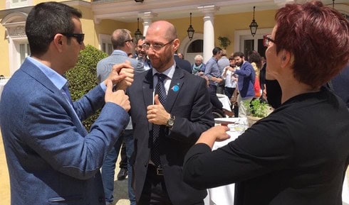 Immagine ravvicinata di Humberto, indossante un abito blu scuro, una camicia bianca e una cravatta blu con puntini bianchi, oltre a una spilla di EUD e EDF, sta comunicando con un'altra persona sordocieca, un uomo con abito blu jeans, camicia celeste, capelli corti neri, pelle chiara e occhiali da sole, utilizzando la lingua dei segni tattile, precisamente segni internazionali. L'altra persona sordocieca, una donna con pelle chiara, capelli corti, occhiali da vista e protesi acustica, indossante una camicia nera, lo guarda attentamente. Alle spalle di Humberto si apre un ambiente all'aperto, con alberi e un locale caratterizzato da colonne, lampade antiche e persone che stanno chiacchierando