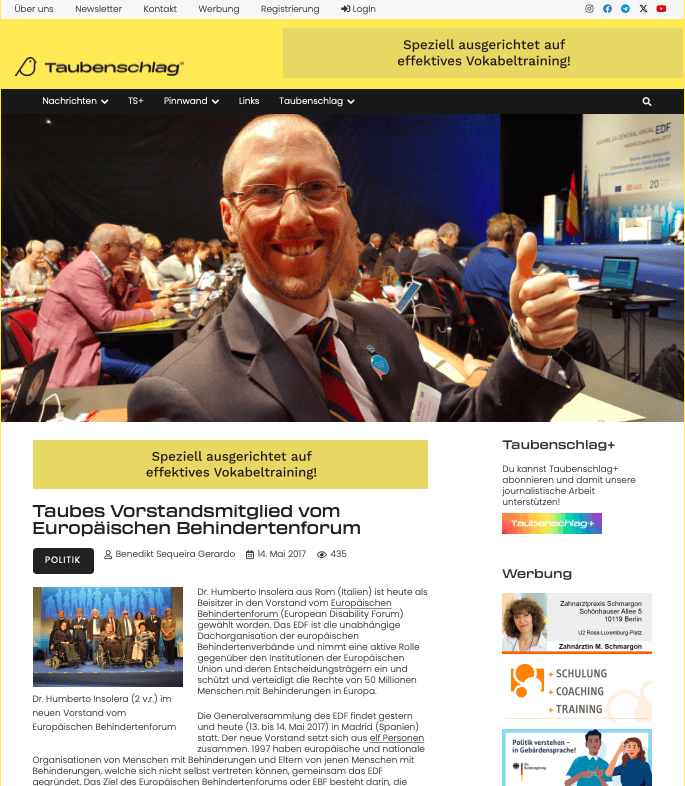 A man in a suit smiles at the camera, giving a thumbs up at a crowded conference hall with visible sign language interpreters in the background.