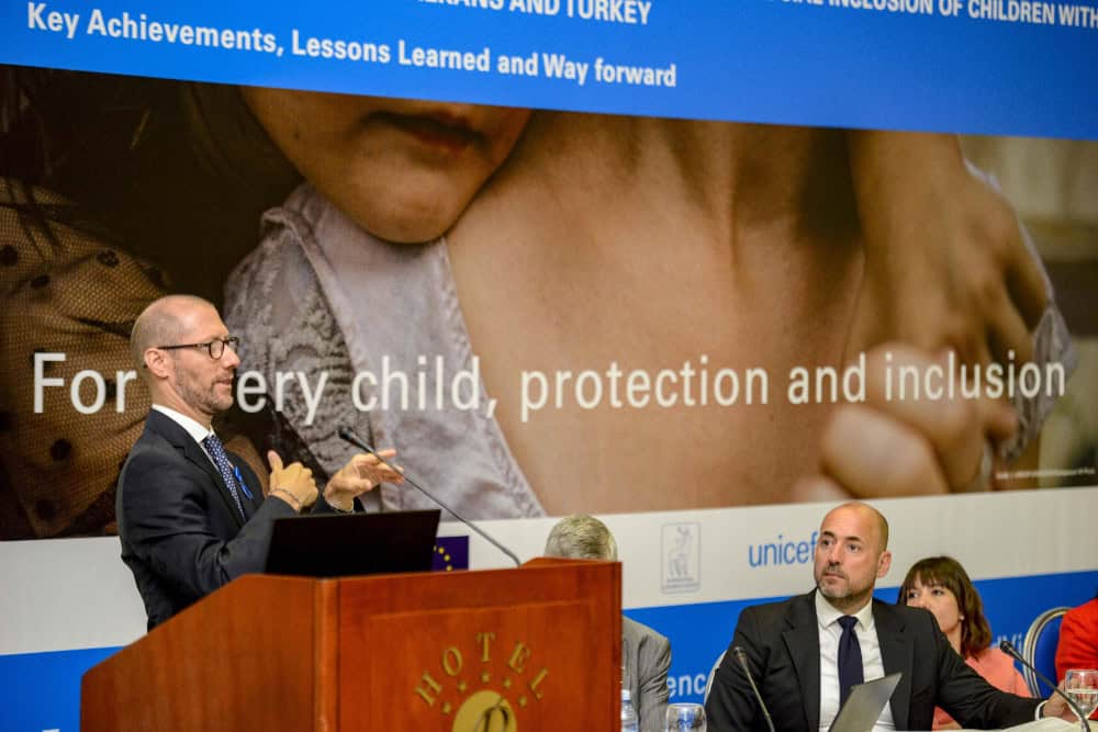 Man delivering a speech at a unicef conference with a banner stating "for every child, protection and inclusion" in the background.